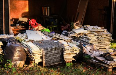 A cluttered outdoor area filled with piles of discarded materials, including cardboard, plastic bags, and various debris. Sunlight casts shadows on the mess.
